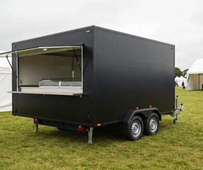 Black twin-axle catering trailer with an open serving hatch at a UK outdoor event