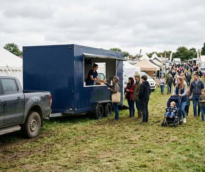 Navy triple axle catering trailer hitched to a 4x4 at a busy UK country show, customers queuing